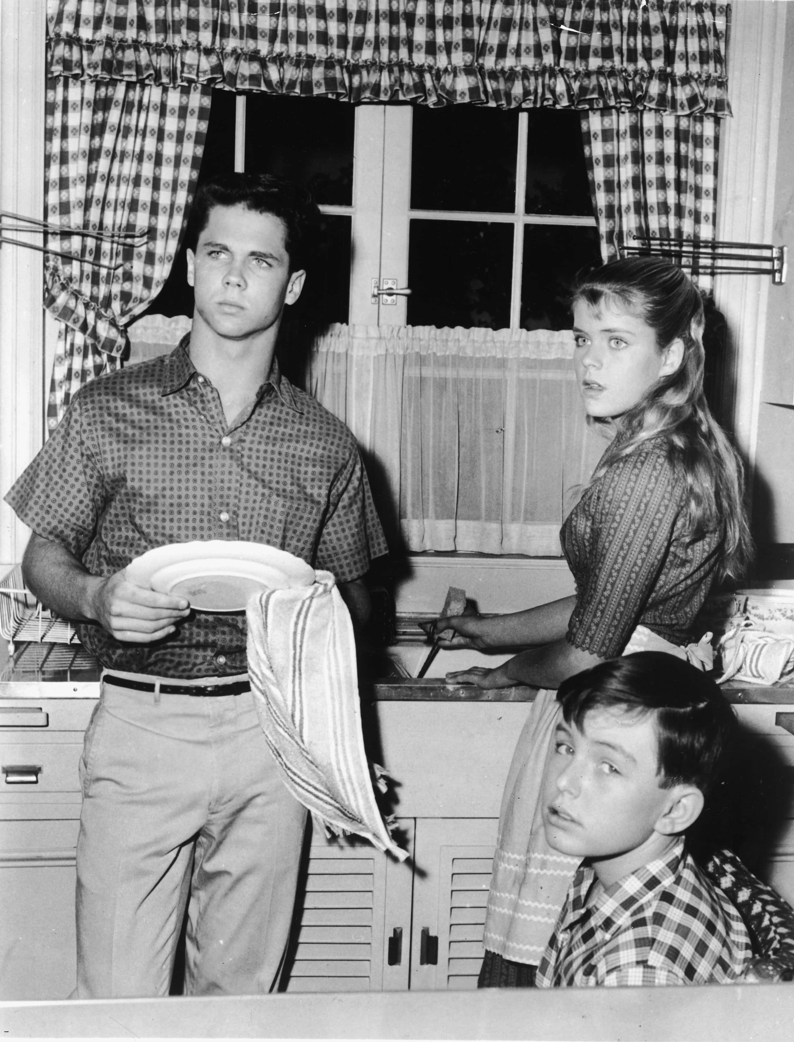 American actor Tony Dow (left, as Wally Cleaver) washes dishes with a towel as he stands in the kitchen alongside an unidentified actress and Jerry Mathers (as the Beaver) who sits and gives the camera a glance, circa 1960. (Photo by ABC Television/Courtesy of Getty Images)